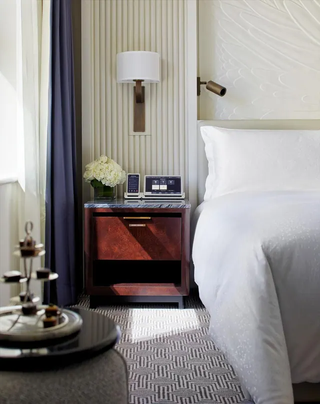 A made white bed sits beside a wooden nightstand with white flowers and a tablet-style control panel, with a tiered tray of pastries on a round table in the foreground.