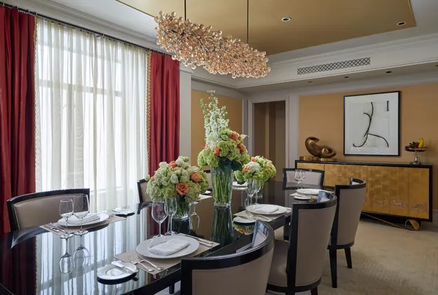 A formally set dining table with white plates, glassware and orange and green flower arrangements, beneath a crystal chandelier, with red curtains at the window and a wooden sideboard against the wall.
