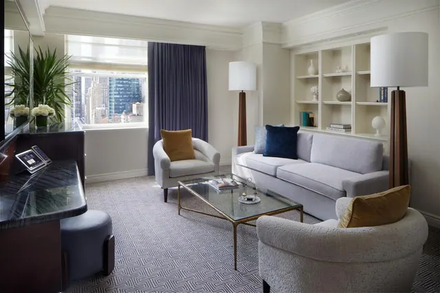 A living area with a grey sofa, two armchairs, a glass coffee table and built-in shelving, with a window showing city buildings.