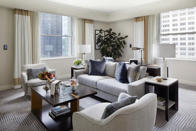 A furnished living room with a grey sofa, armchairs, a coffee table and large windows overlooking high-rise buildings.