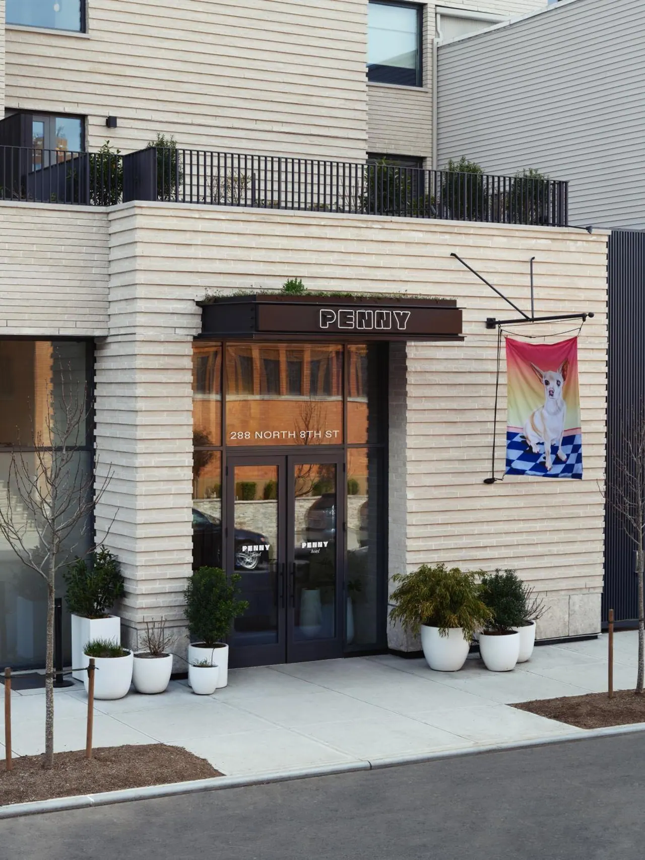 The entrance of a light-brick building with a dark PENNY sign above glass doors, white planters on the sidewalk, and a hanging banner with a dog print on the right.