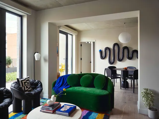 A living area with a green velvet curved sofa, black leather armchairs and a round coffee table, with a dining table visible through an archway.