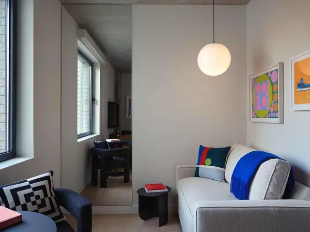 A living area with a light grey sofa, a blue throw and cushions, a round white pendant lamp, framed artwork on the wall, and a mirrored panel reflecting a bedroom with a dark armchair.
