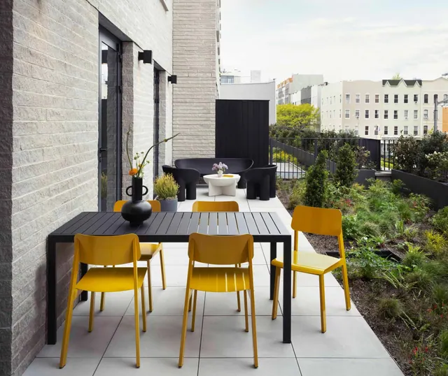 A paved terrace with a black dining table, yellow chairs, a black sofa and a low white coffee table, with a planted bed and low-rise residential buildings beyond the railing.