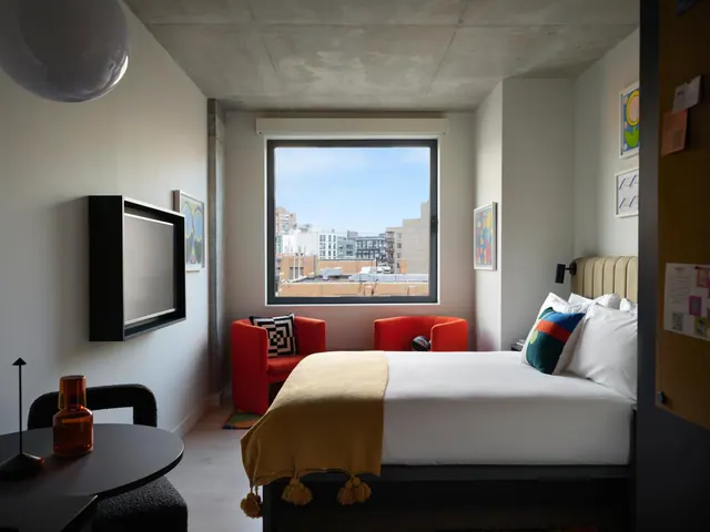 A hotel bedroom with a double bed, two red armchairs, a wall-mounted television and a window looking out over city buildings.