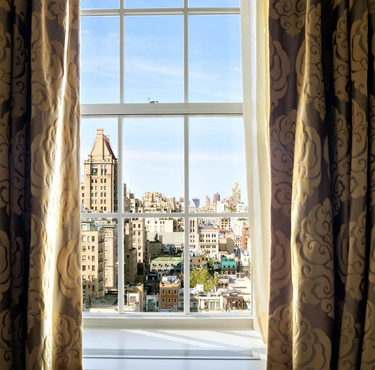 A tall window with patterned curtains looks out over a dense cluster of city buildings and rooftops under a clear blue sky.