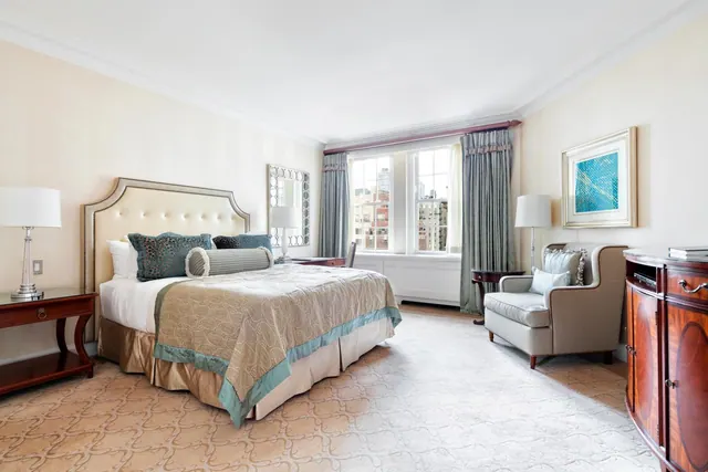 A bedroom with a king bed, bedside lamps, an armchair, a wooden cabinet and windows looking onto nearby city buildings.
