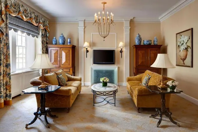 A formal living area with two gold upholstered sofas facing each other across a round coffee table, a wall-mounted television above a fireplace, two wooden cabinets with blue and white vases, and a chandelier overhead.