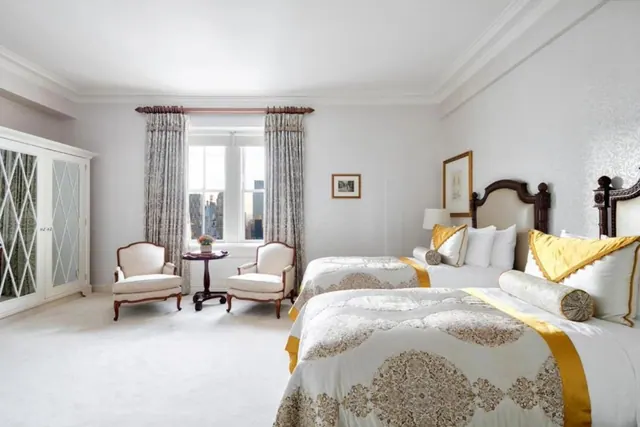 A bedroom with two twin beds with gold-trimmed bedding, two armchairs and a small round table beside a window showing city buildings.