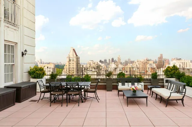 A rooftop terrace with a dining table, chairs and lounge seating overlooks a city skyline under a partly cloudy sky.