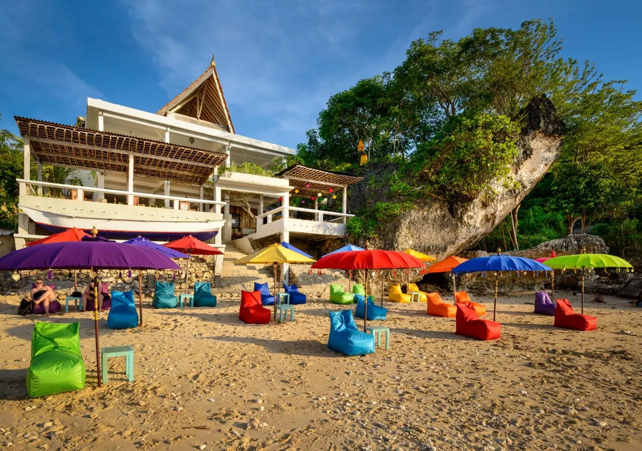 Colourful bean bags and parasols are arranged on a sandy beach in front of a white two-storey building with a thatched roof and a rocky outcrop behind it.