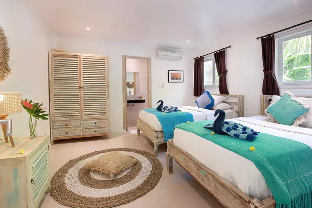 A bedroom with two beds dressed in teal throws, a wooden wardrobe, a round woven rug, and a glimpse of an en suite bathroom through an open doorway.