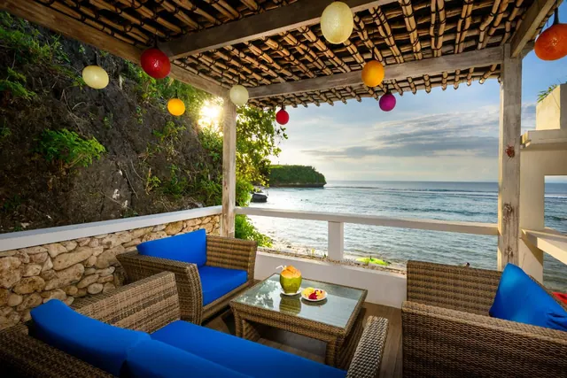 A covered terrace with blue-cushioned wicker seating and a table holding a coconut drink and fruit, overlooking the sea.