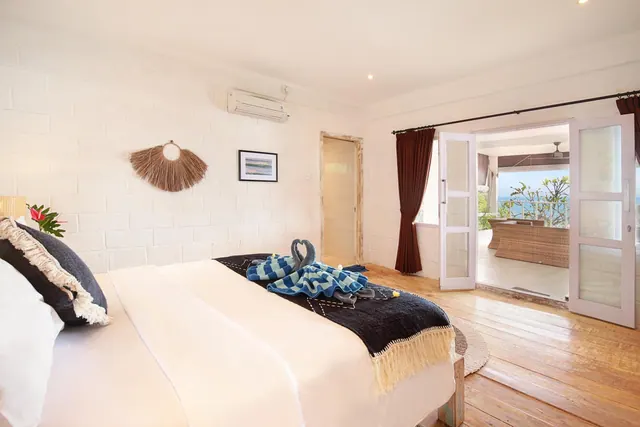 A made-up bed with a towel swan on a dark throw sits in a white-walled bedroom, with open doors onto a balcony overlooking the sea.