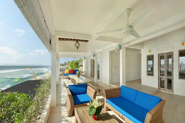 A covered terrace with rattan sofas, blue cushions and a ceiling fan, with the ocean and breaking waves visible to the left.