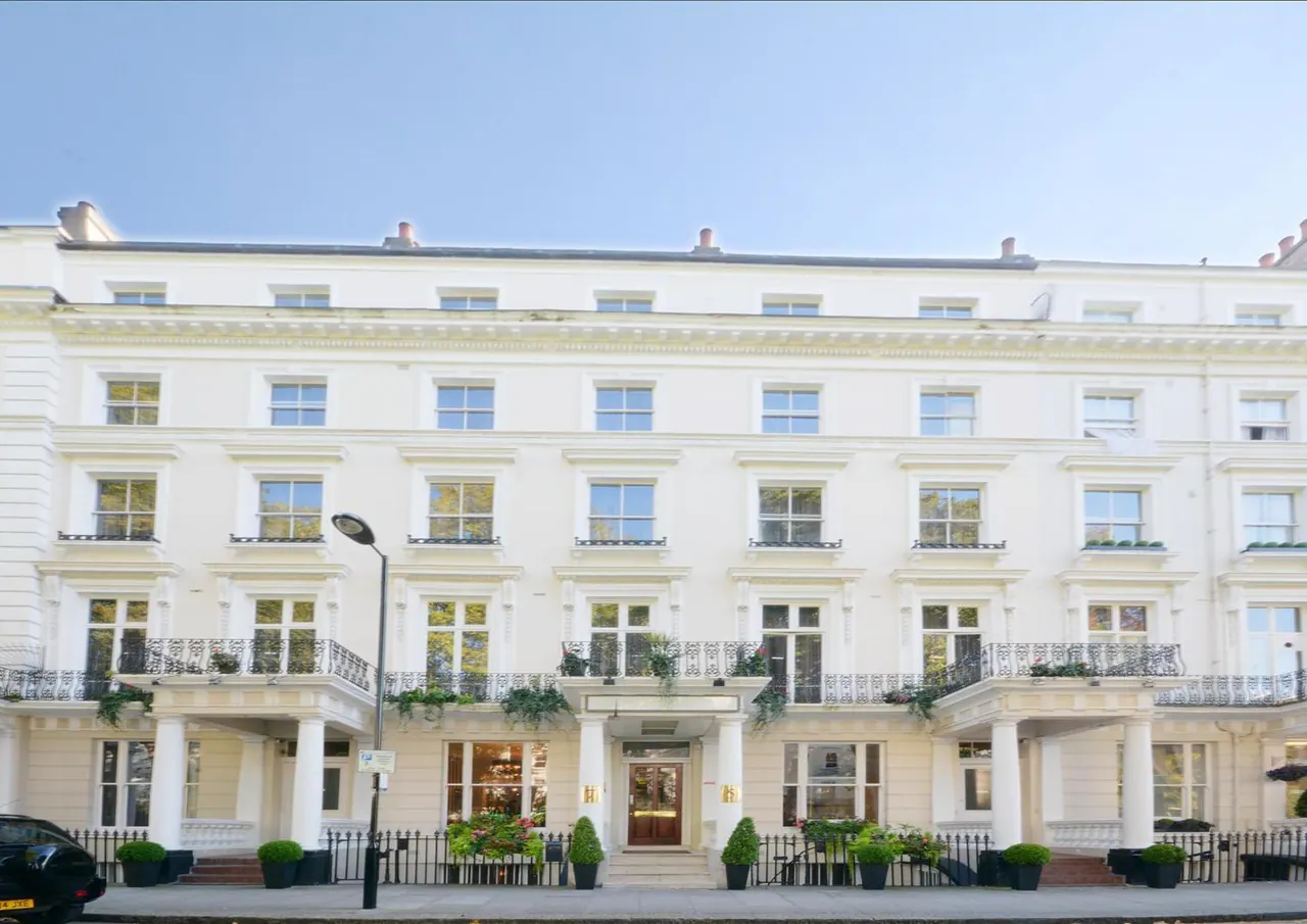 The white stucco facade of a multi-storey terraced building with a central entrance, columns, planters and iron railings is shown, with a street lamp and a dark car at the left edge.