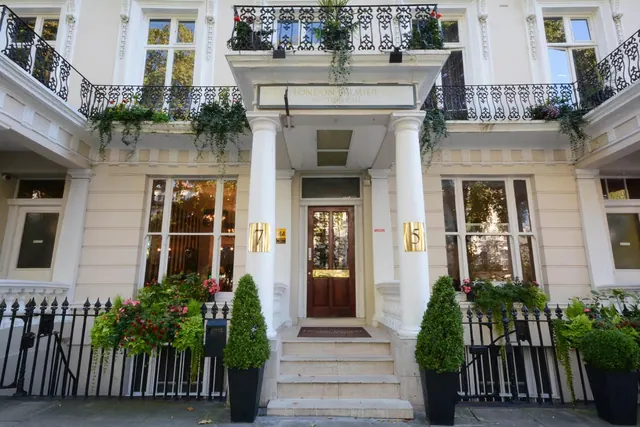 The cream stucco front of the London Premier Notting Hill hotel has black iron railings, potted topiary and a central entrance with stone steps and a wooden door.