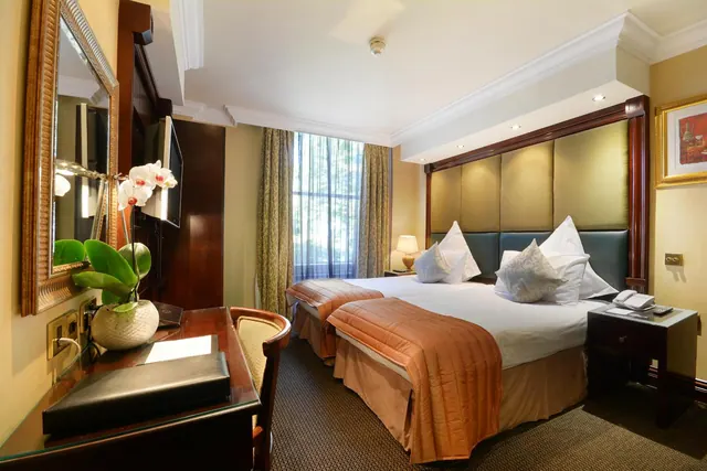 A hotel bedroom with two beds under orange throws, a wooden desk with an orchid plant and a telephone, and a window with patterned curtains.