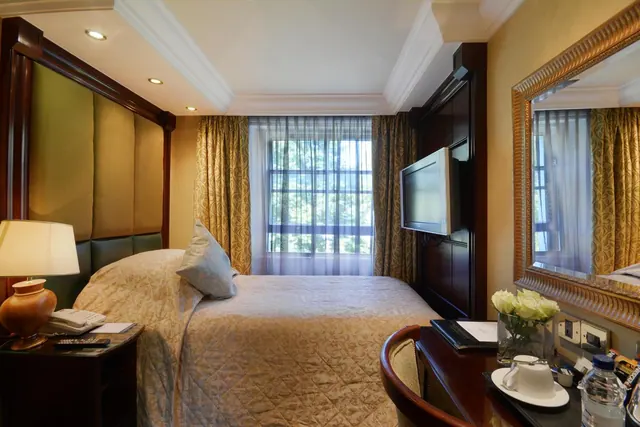 A double bed with gold quilted bedding sits in a hotel bedroom beside a window with sheer curtains, a wall-mounted television, and a desk holding a flower vase and a cup on a saucer.