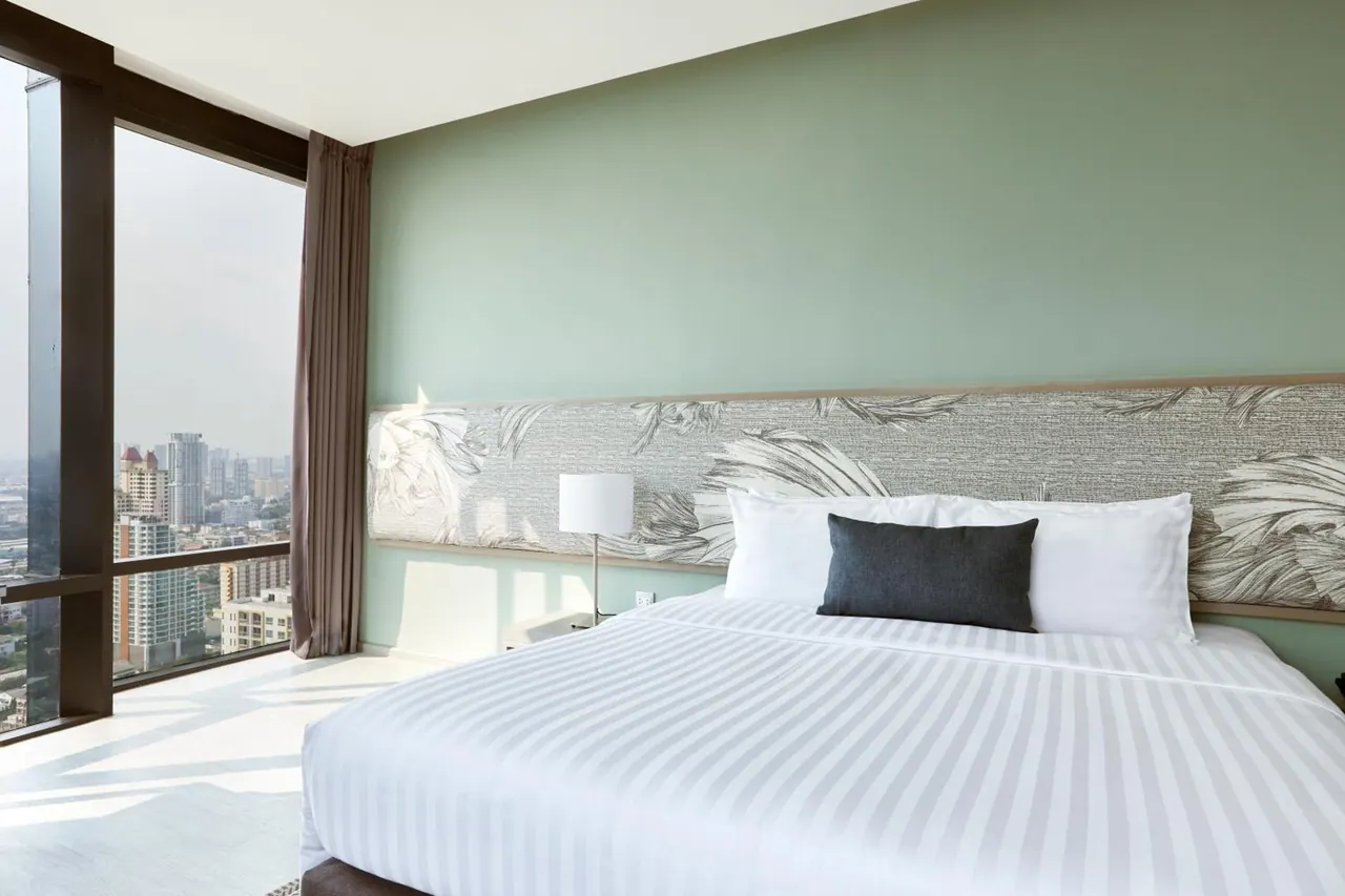 A made king bed with striped white linen and a dark cushion sits beside floor-to-ceiling windows overlooking a hazy high-rise cityscape.