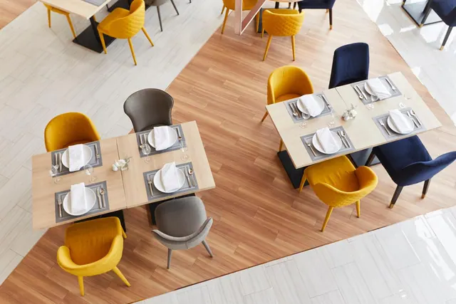 Overhead view of a dining room with several tables set with plates, napkins and cutlery, and yellow, grey and navy chairs on a wood-effect floor.
