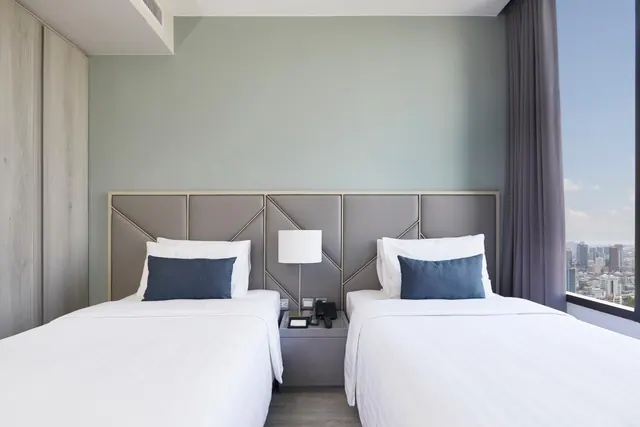 A bedroom with two single beds with white linen and blue cushions, a grey padded headboard, a bedside lamp and telephone, and a tall window showing city buildings.