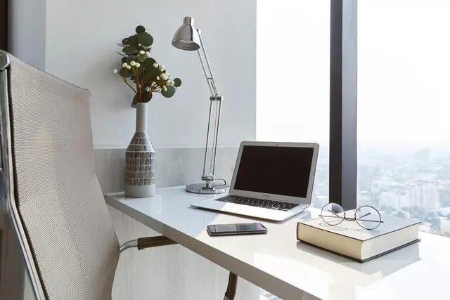 A white desk by a floor-to-ceiling window holds a laptop, a desk lamp, a vase of foliage, a phone, glasses and a book, with a city view outside.