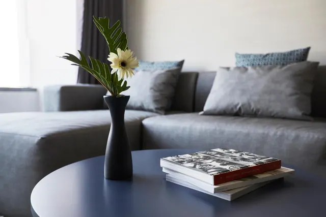 A grey sofa with cushions sits behind a round navy coffee table holding a black vase with a gerbera flower and a stack of books.
