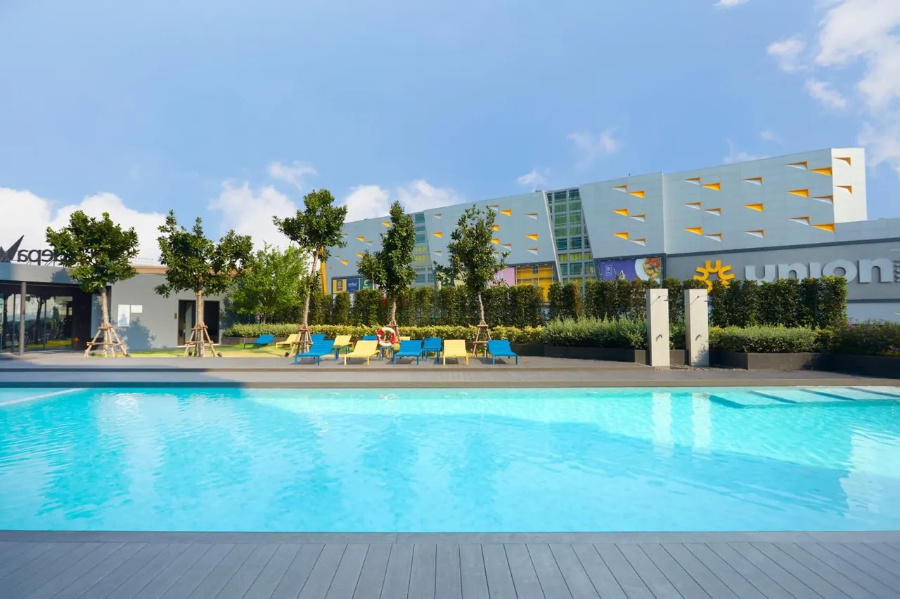 An outdoor swimming pool in the foreground with blue and yellow sun loungers, trees and hedges, and a multi-storey building with a union sign behind it.