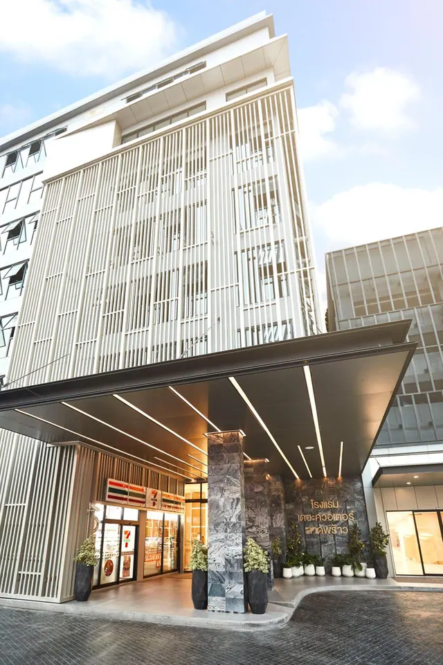 Low-angle view of a white multi-storey hotel building with a covered entrance canopy, marble columns, potted plants and a convenience store at the left.