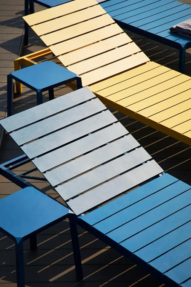 Yellow, grey and blue sun loungers and small side tables sit on a dark wooden deck in strong sunlight.