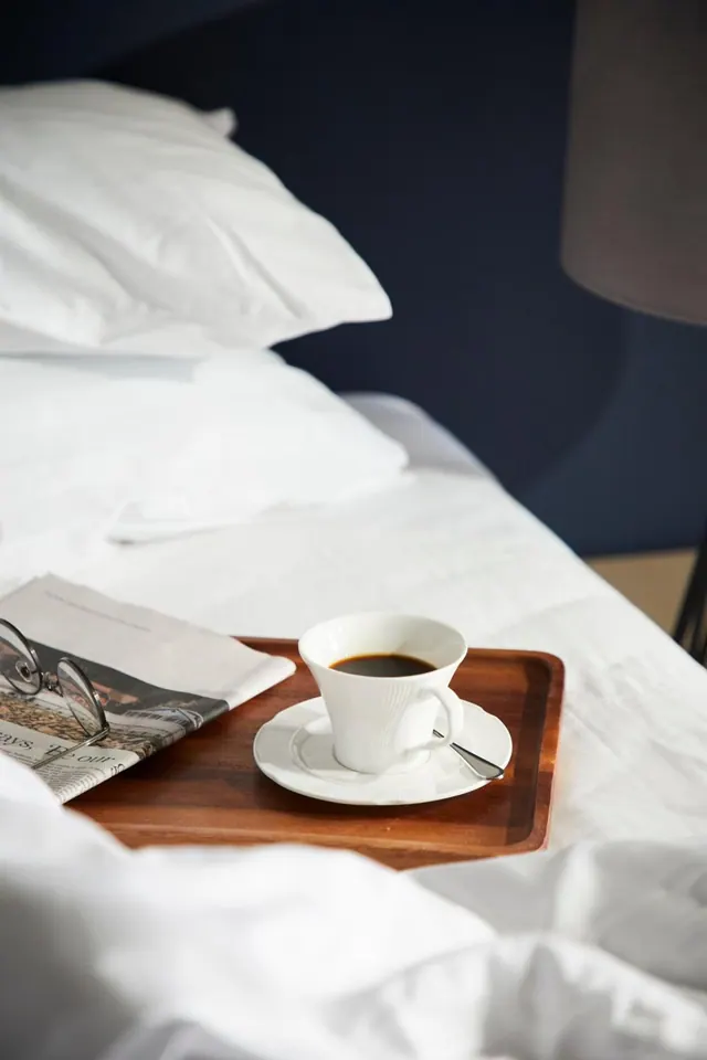 A white bed with pillows and white linen has a wooden tray holding a cup of black coffee on a saucer, a folded newspaper and reading glasses.