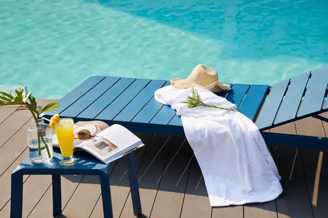 A blue sun lounger beside the pool holds a white towel and straw hat, with a side table nearby holding orange juice, a magazine, and sunglasses.