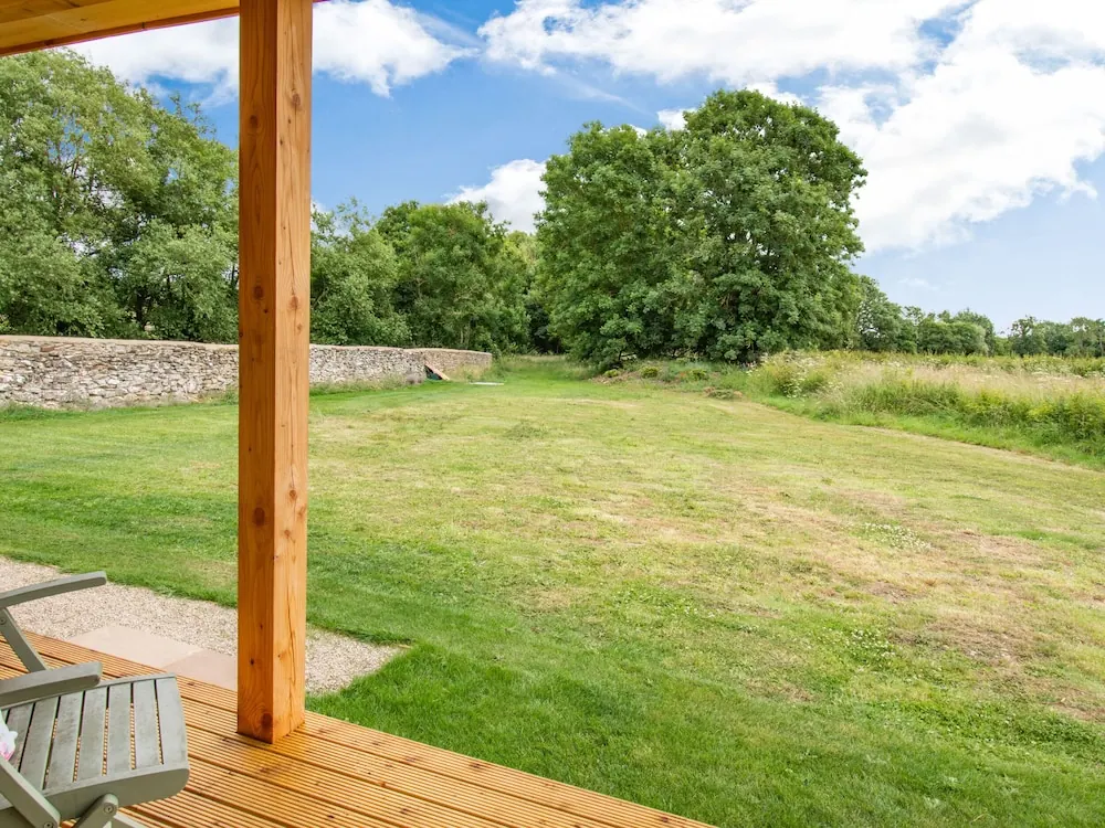 A covered wooden deck with a grey chair looks out over a mown lawn, a line of trees and a stone wall.