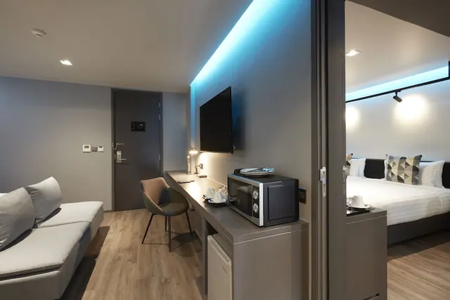 A hotel room living area with a grey sofa, a desk holding a microwave above a mini fridge, a wall-mounted TV with blue LED lighting, and a double bed visible through the doorway on the right.