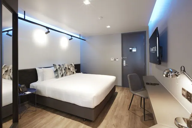 A modern hotel bedroom with a king bed, a wall-mounted television, a work desk with a chair, and a door to the corridor.