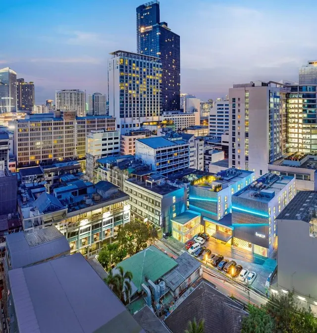 Elevated dusk view over a dense Bangkok cityscape with high-rise towers, a lit street with parked cars, and a hotel building with blue accent lighting.
