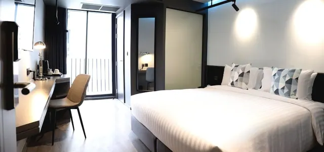 A modern hotel bedroom with a king bed, a desk with a lamp and chair, and a glass sliding door leading to a balcony.