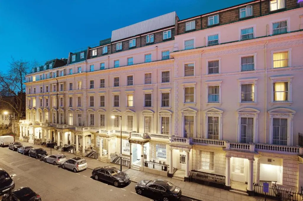 A long white stucco terrace building with lit windows and balconies at dusk, with cars parked along the street in front.