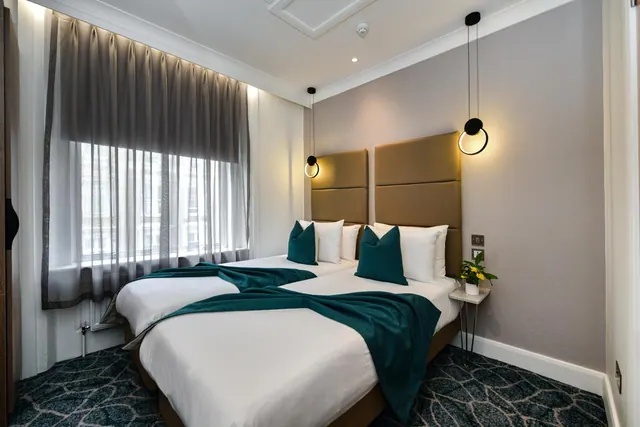 A bedroom with two beds side by side under white bedding, teal throws and cushions, tall padded headboards, and a pendant light on each side, with a sheer-curtained window on the left.