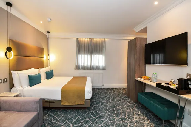 A double bed with teal cushions, a wall-mounted television, a kettle on a tray at a desk, and a grey sofa in the foreground of a hotel bedroom.