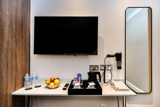 A wall-mounted television, switched off, sits above a table holding water bottles, a fruit bowl, a remote control, a tea tray with a kettle, and a corded telephone, beside a tall mirror and a wall-mounted hairdryer.