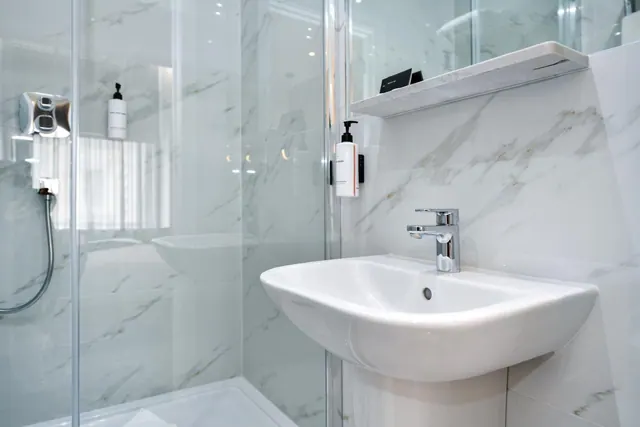 A bathroom with a glass-walled walk-in shower, marble-effect wall panels, a pedestal sink with a chrome tap, and wall-mounted toiletry dispensers.