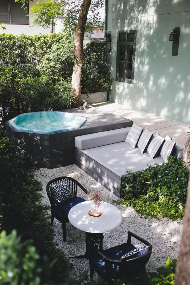 A private garden patio with a hot tub, a grey daybed with striped cushions, and a small table with two chairs on gravel.
