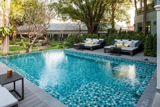 A mosaic-tiled swimming pool with grey daybeds and cushions on the deck, with trees and a traditional tiled-roof building behind.