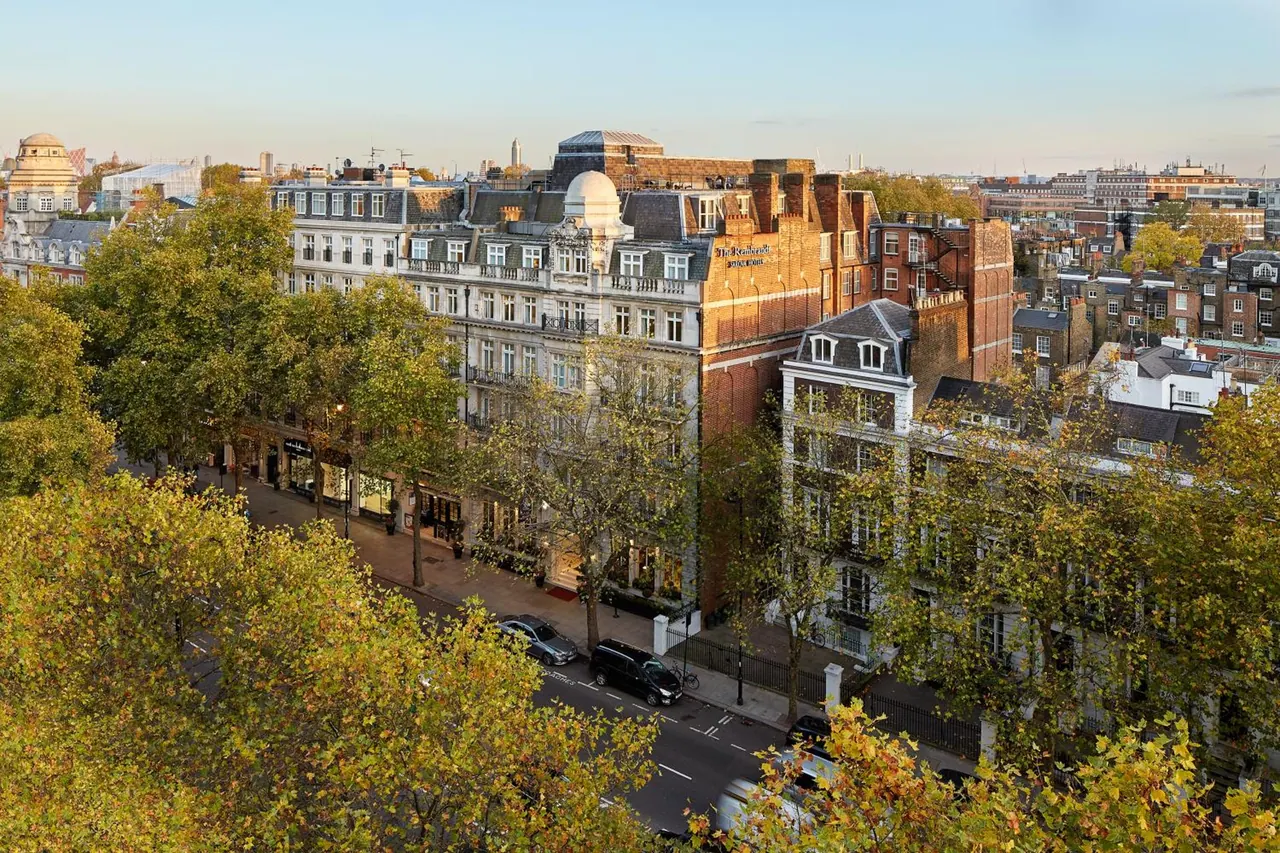 Elevated view down a tree-lined London street with ornate cream and brick hotel buildings, parked cars and rooftops in the distance.
