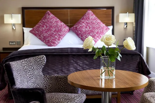 A double bed with pink cushions and a purple throw sits beside a leopard-print armchair and a round wooden table holding a vase of white roses.