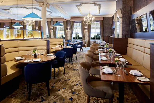 A restaurant dining room with laid tables, blue upholstered chairs, banquette seating and chandeliers under an ornate plaster ceiling.