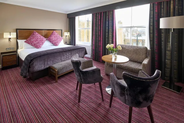 A hotel bedroom with a king bed and pink cushions, a round table with two armchairs, a leopard-print armchair, and windows looking onto neighbouring brick buildings.