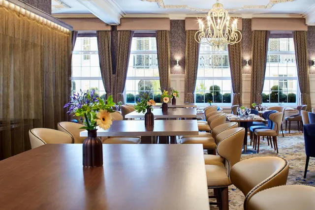 A restaurant dining room with set tables, tan leather chairs, a chandelier and tall windows looking onto neighbouring buildings.